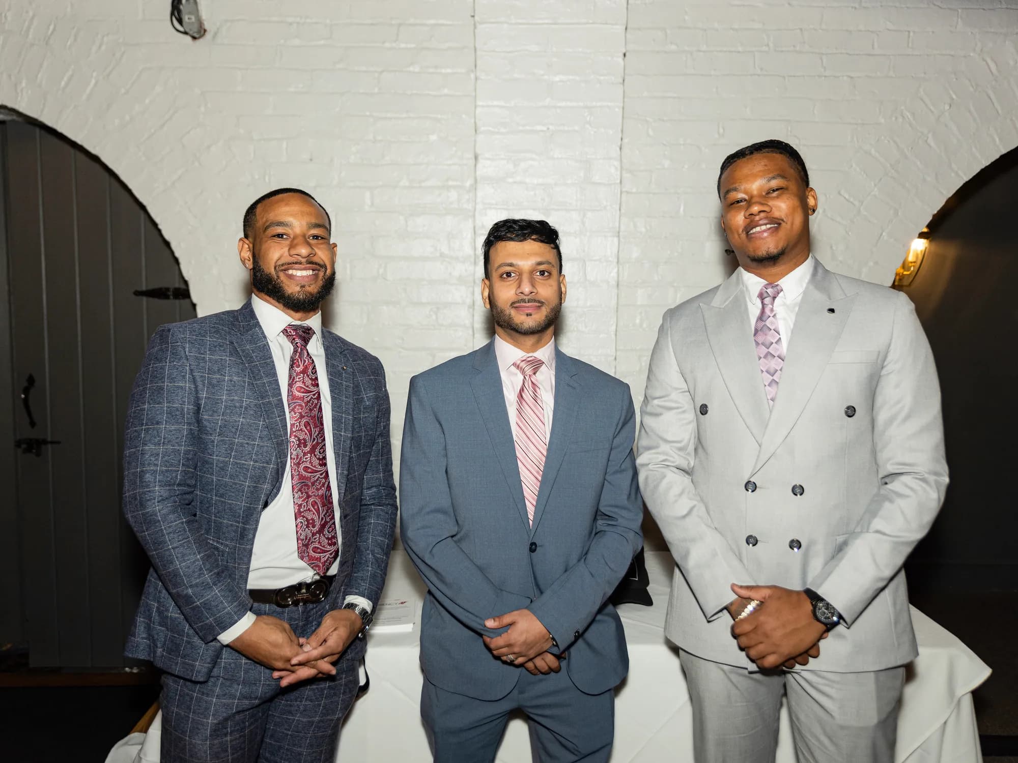 Three guests in suits standing together against a white brick wall at an event.