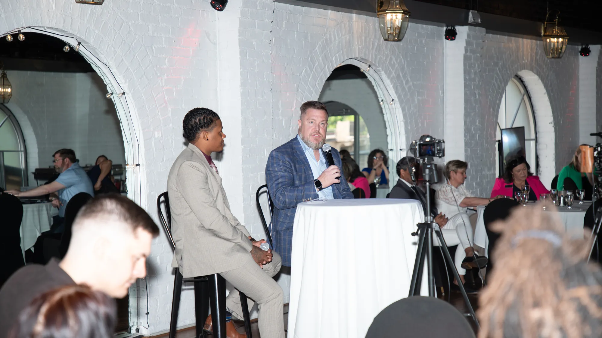 A live panel mid-conversation, one speaker on a microphone, a camera on a tripod filming it and a seated audience watching.