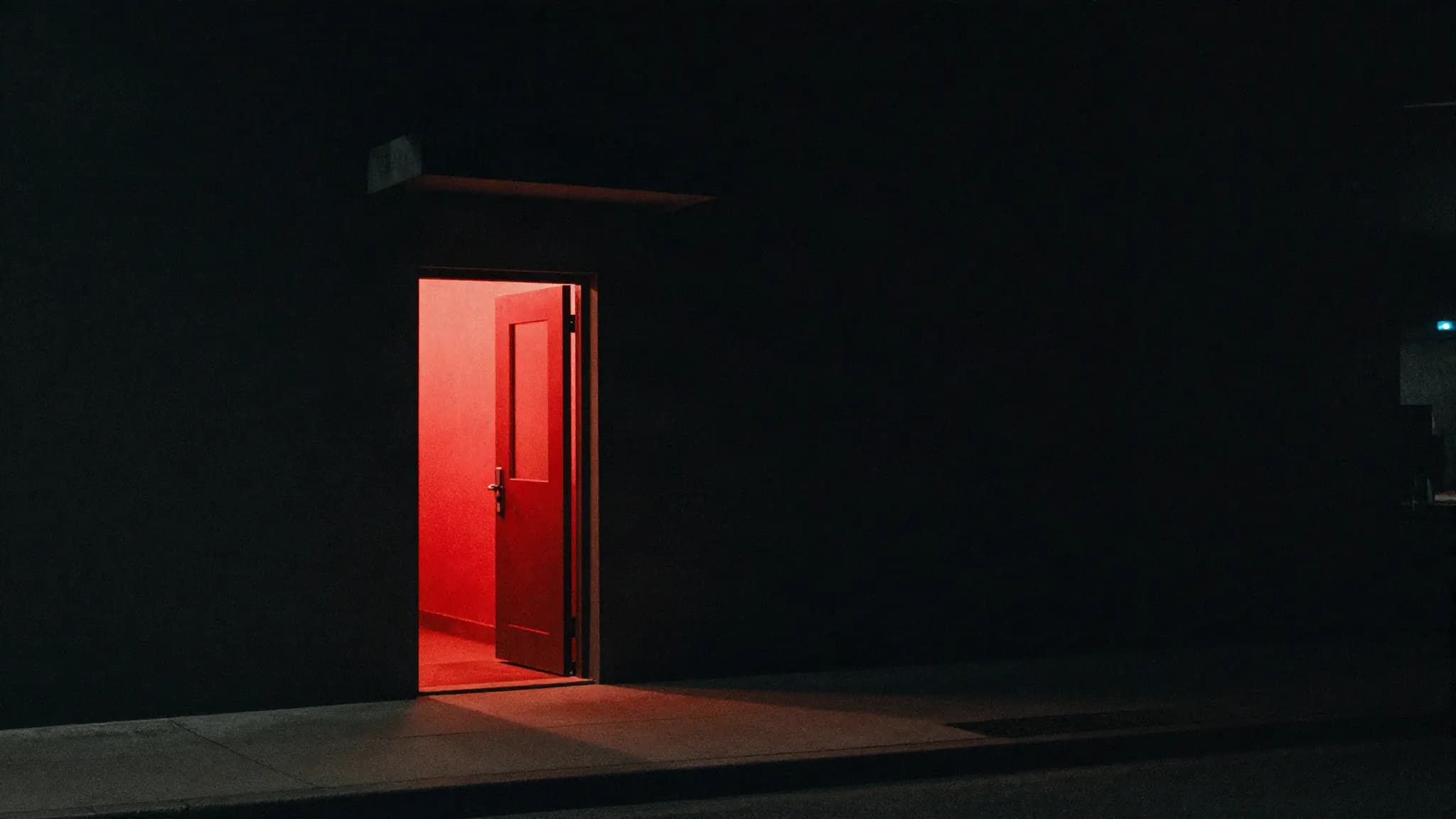 A single lit doorway in a dark building facade at night, warm and red light spilling across the sidewalk.