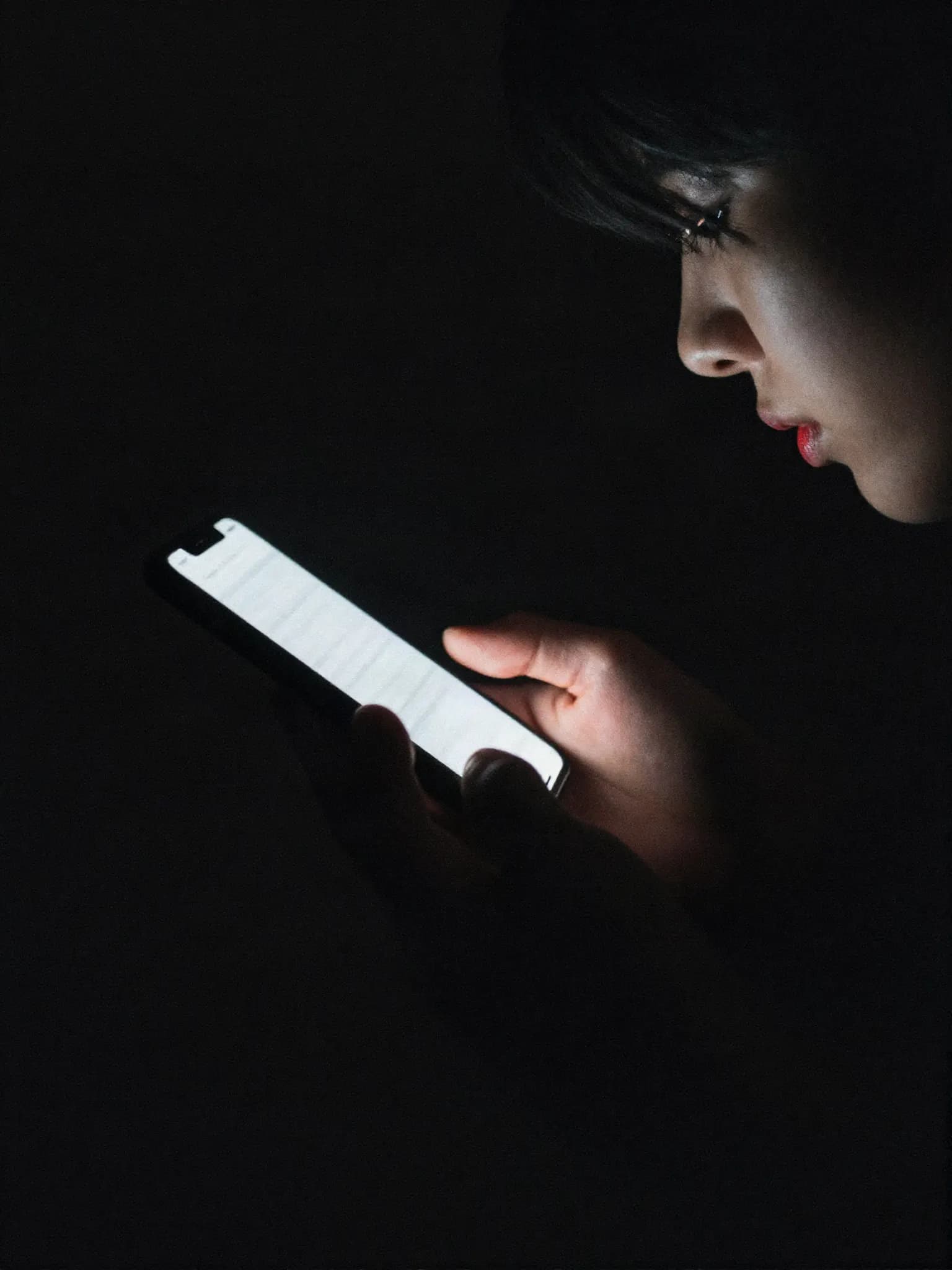 A hand holding a phone in a dark room, the screen throwing hard light across the knuckles.