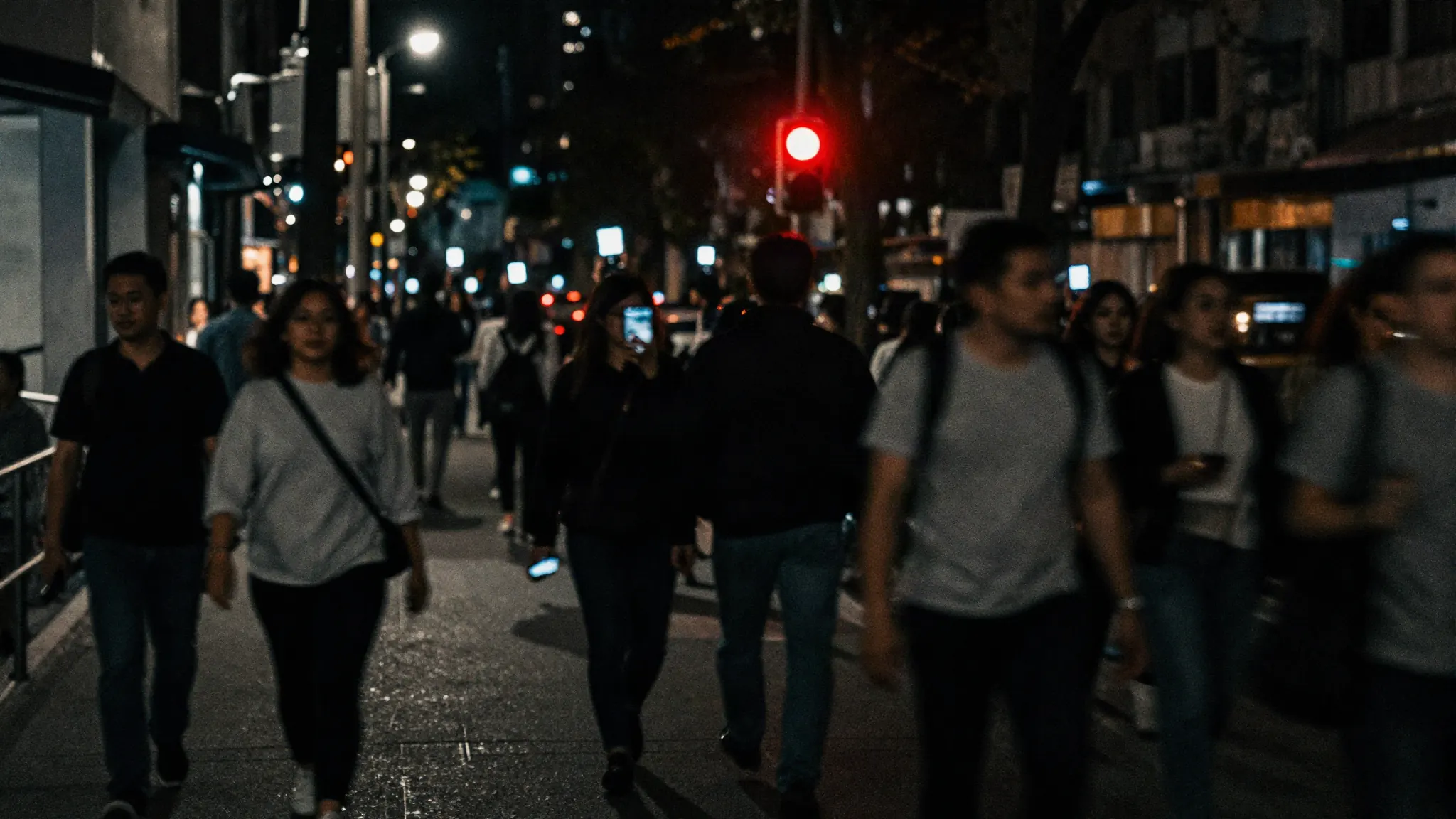 A crowded night sidewalk shot on a long exposure, bodies smeared into streaks and phone screens scattered through the blur like points of light.