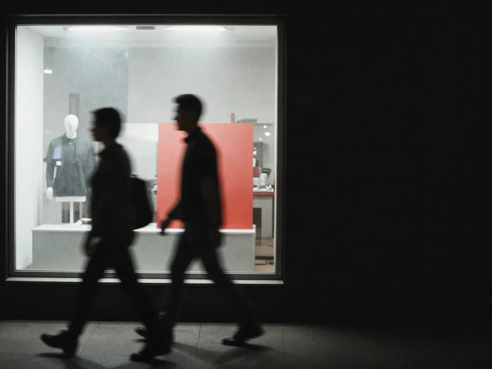 A badly motion-blurred figure walking past a brightly lit shop window at night.