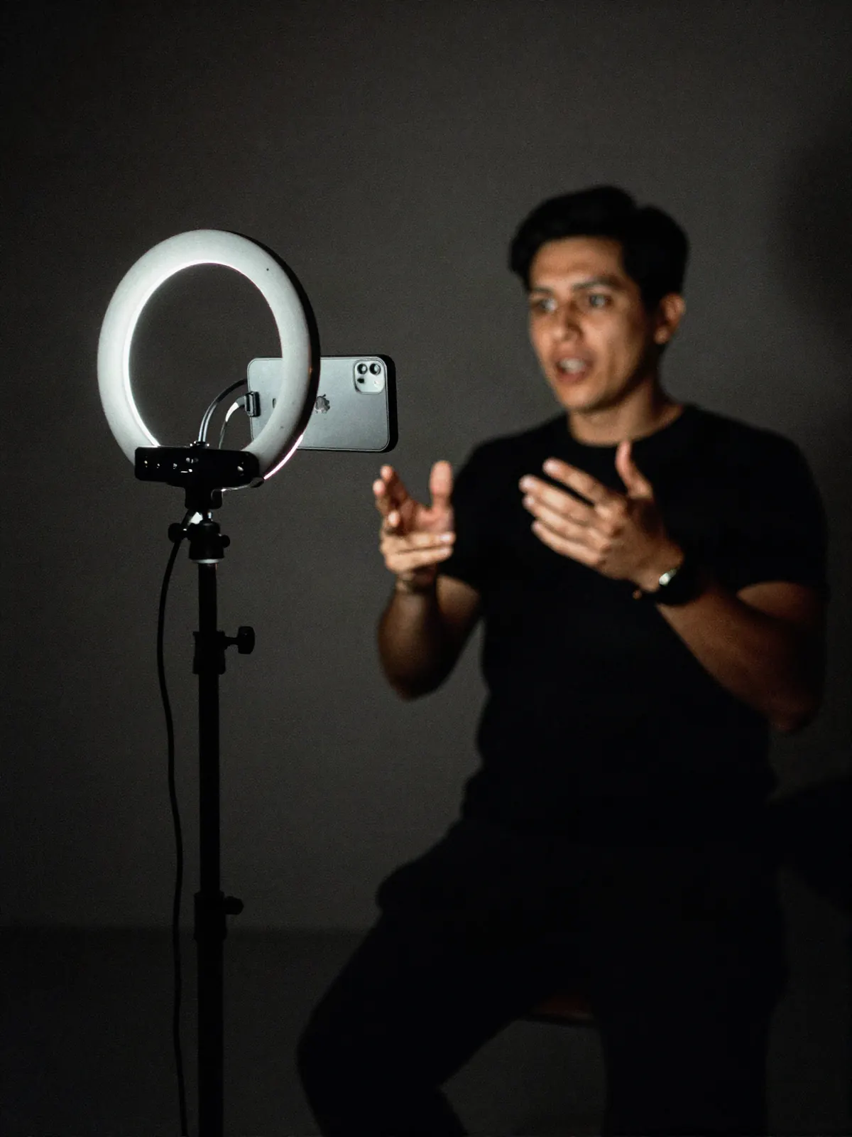 A phone clamped to a tripod beside a ring light, with a person gesturing far out of focus behind it.
