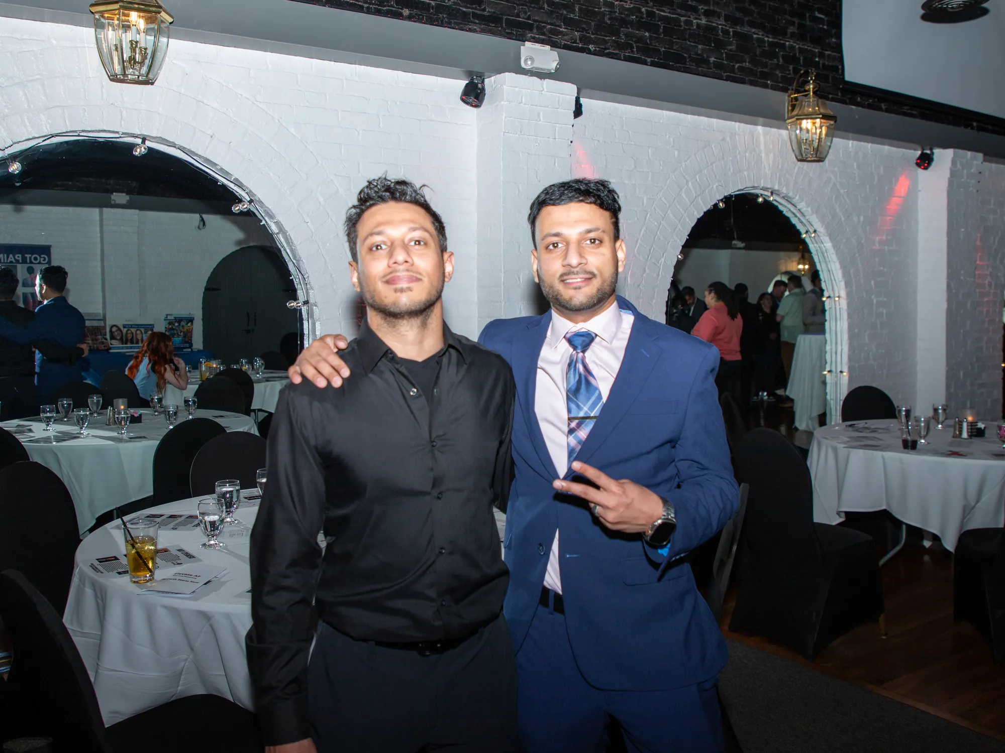 Two guests on the floor of an event, the room of set tables and arched brickwork behind them.