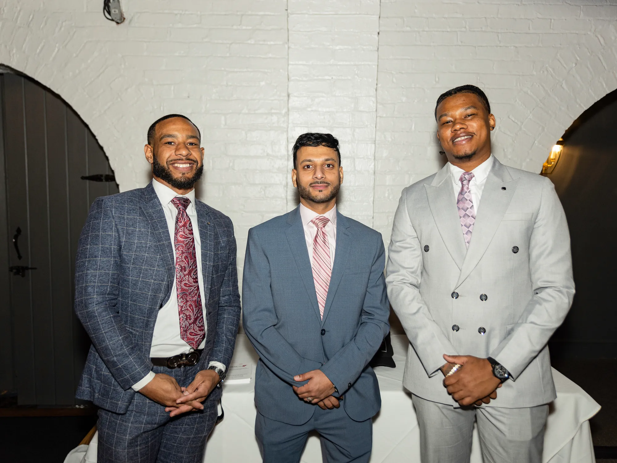 Three guests in suits standing together against a white brick wall at an event.
