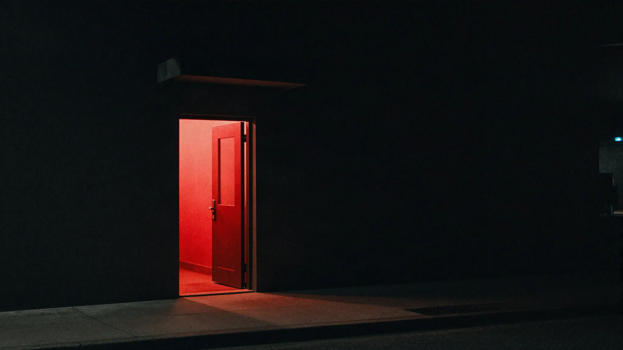 A single lit doorway in a dark building facade at night, warm and red light spilling across the sidewalk.