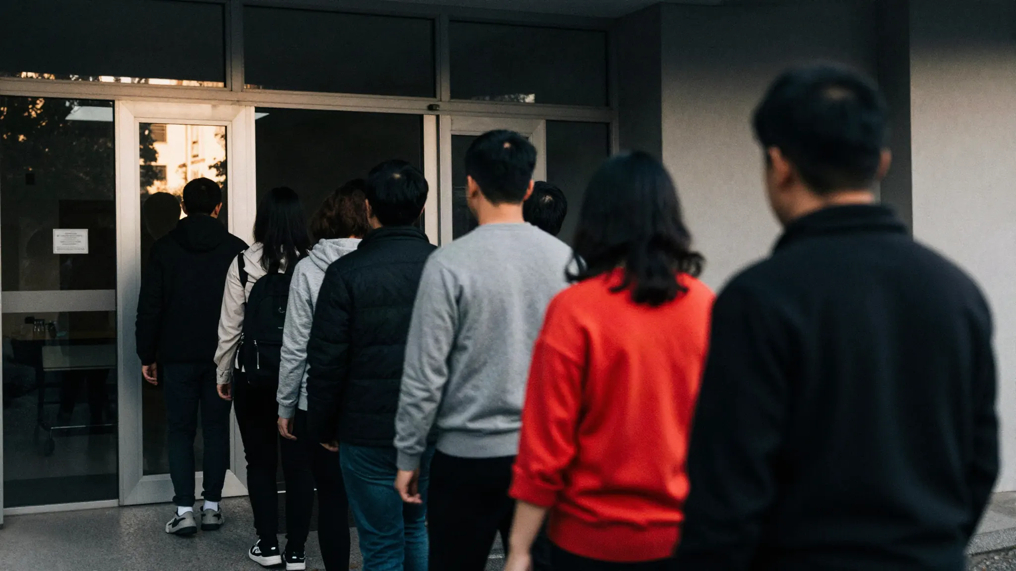 People filing through the entrance of a business in the morning, sunlight flaring off the glass door.