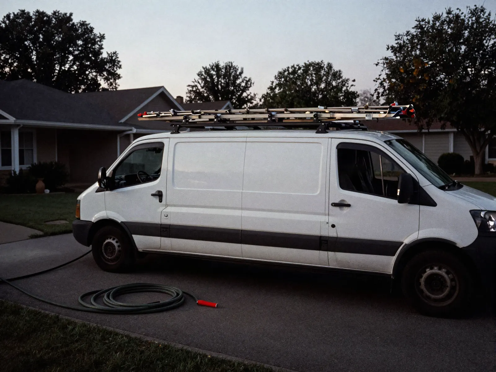 A work van backed into a suburban driveway at first light with a ladder on the roof rack and tools on the pavement.