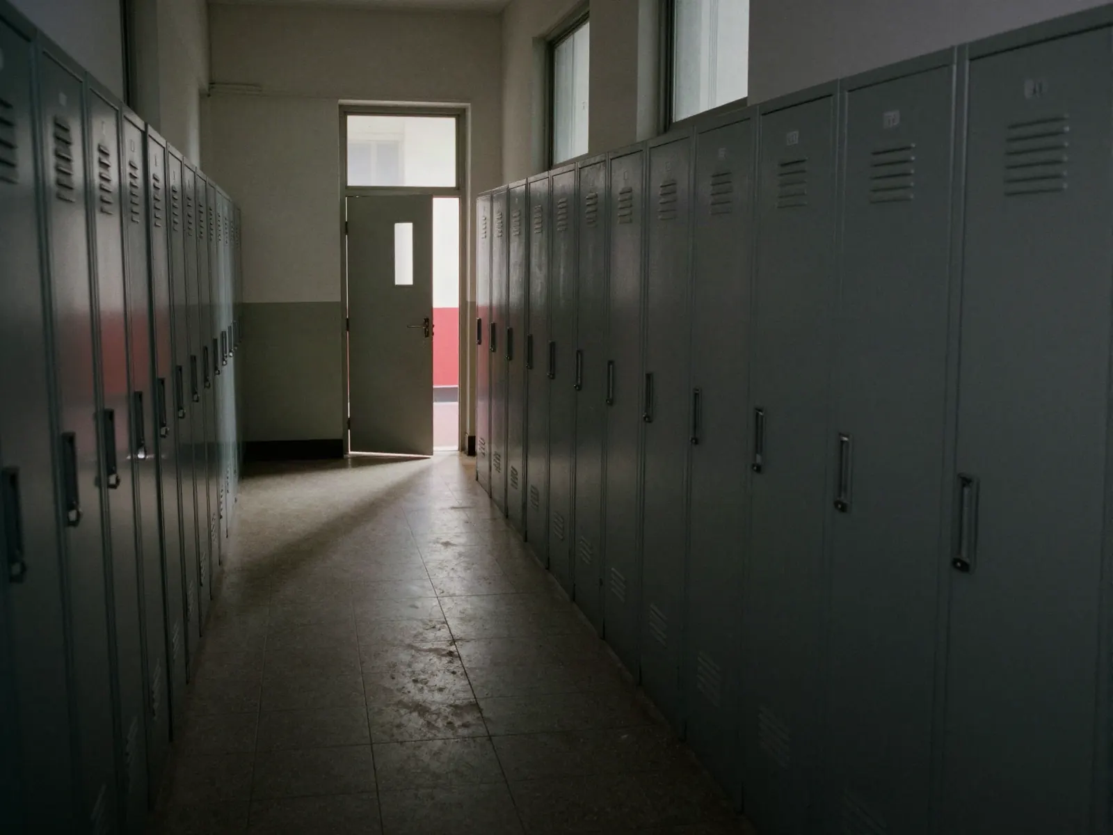An empty school corridor with banks of lockers and hard shafts of window light across a worn tile floor.